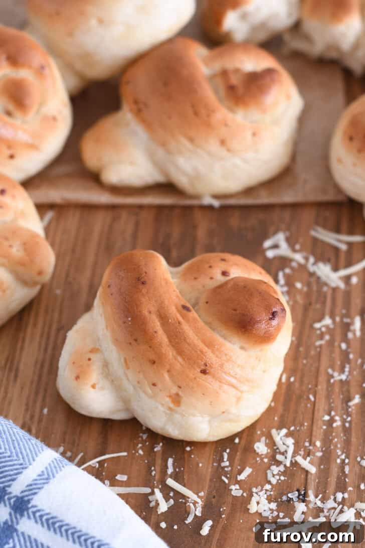 Simple Parmesan Garlic Knots 2 A freshly baked Parmesan breadstick knot, garnished with shredded Parmesan cheese and resting on a rustic wooden board beside a white and blue patterned cloth.