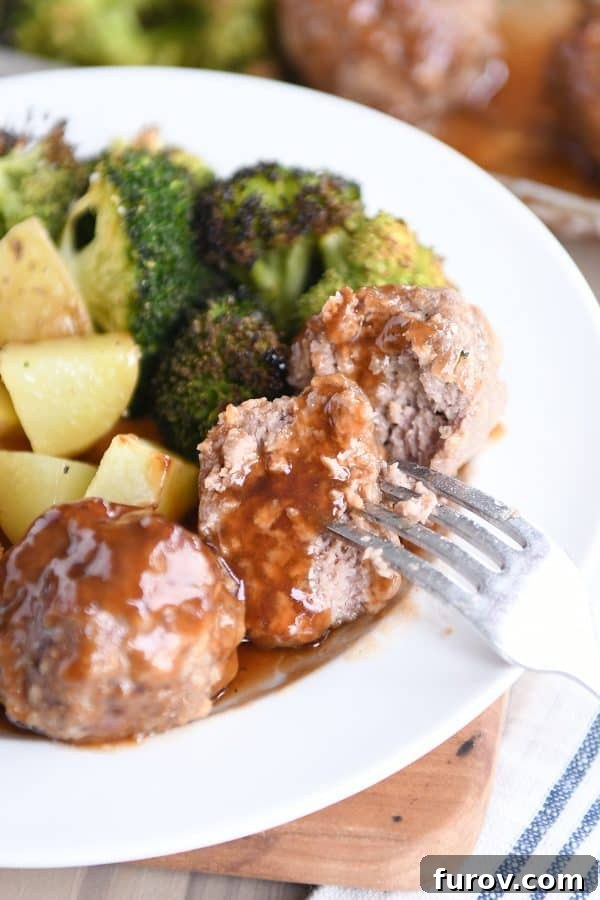 One-Pan Sweet & Sour Meatballs, Roasted Potatoes & Broccoli 4 A fork piercing half of a perfectly cooked sweet and sour sheet pan meatball, resting on a white plate, showcasing its tender interior and glazed exterior.