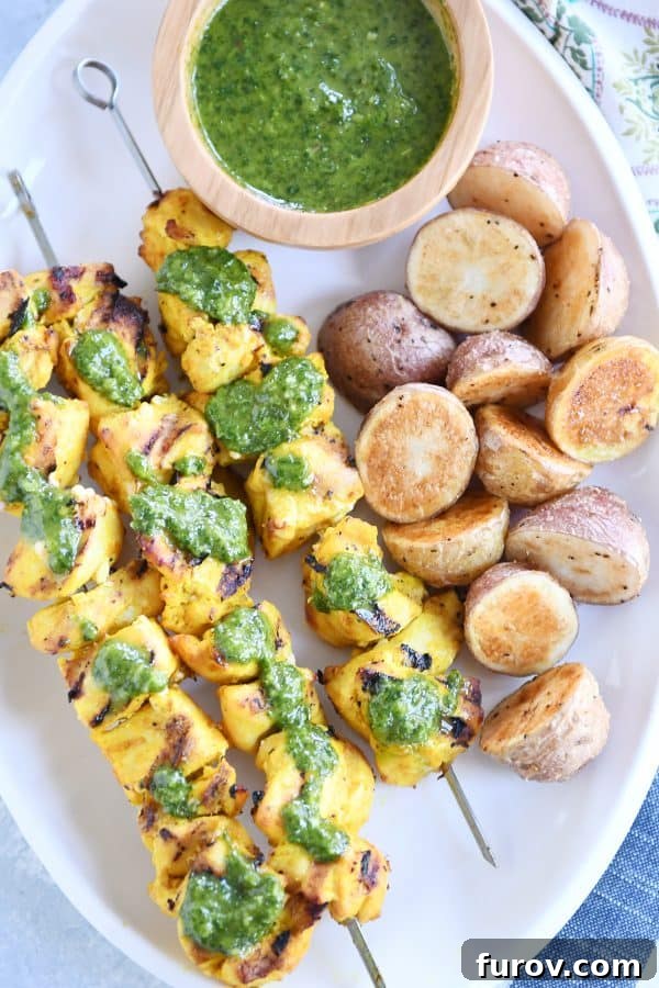 Moroccan Chicken Skewers with Zesty Green Sauce 5 Top-down view of a white platter showcasing Moroccan chicken skewers, a bowl of green sauce, and roasted potatoes.