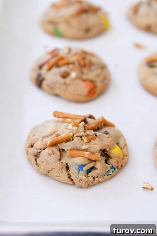 Sweet and Salty Peanut Butter Kitchen Sink Cookies 3 A perfectly baked peanut butter kitchen sink cookie rests on white parchment paper, highlighting its golden edges and studded surface.