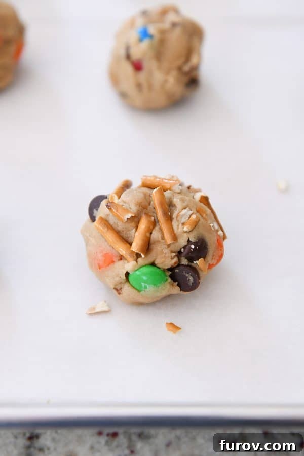 Sweet and Salty Peanut Butter Kitchen Sink Cookies 4 Unbaked peanut butter kitchen sink cookie dough balls, studded with various inclusions, arranged on a baking tray lined with parchment paper.