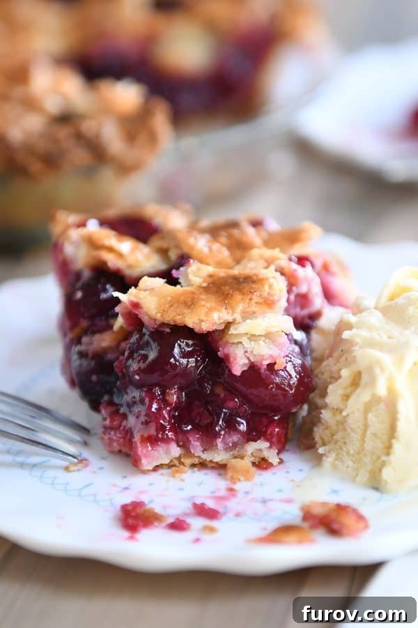 A golden-crusted cherry pie with a slice removed, revealing the vibrant red filling. A scoop of vanilla ice cream rests beside it.