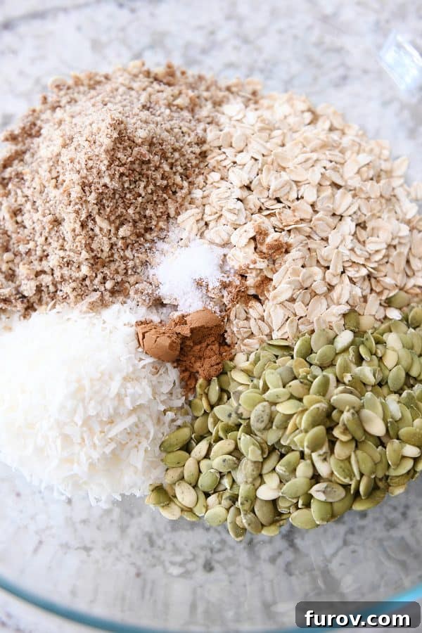 Glass bowl with ingredients for granola: coconut, pepitas, cinnamon, oats, pecans.