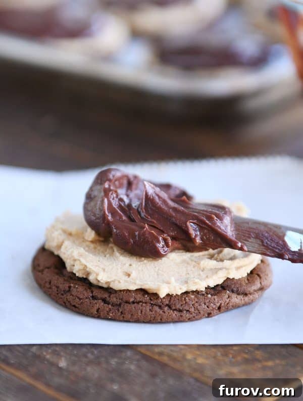 A hand gently spreading chocolate ganache over a peanut butter frosted chocolate cookie.