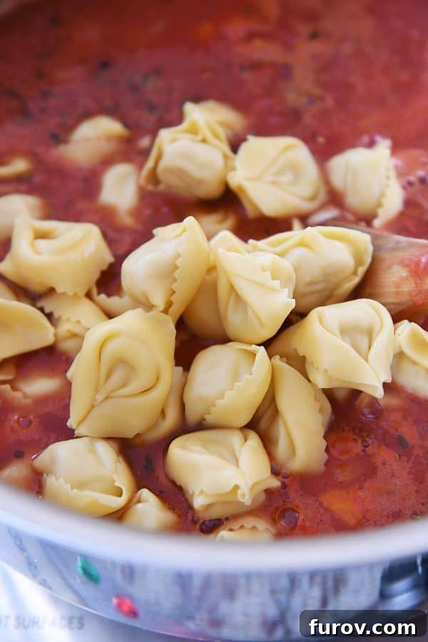 A close-up shot shows tender cheese tortellini being added to the simmering Tuscan tortellini soup, ready to absorb the rich flavors.
