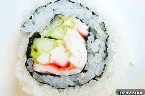 A perfectly rolled California sushi roll, viewed from the side, revealing its layers of rice, nori, avocado, cucumber, and crab. The rice-out construction is clearly visible.