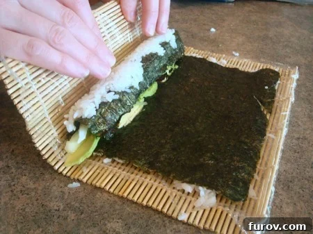 The bamboo mat is lifted and repositioned above the partially rolled sushi, preparing for the next push to tighten the roll.
