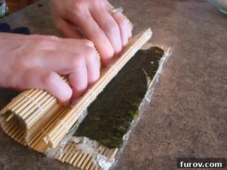 A hand pressing down on the bamboo mat, which is wrapped around the nearly complete California sushi roll, showcasing the final tightening.
