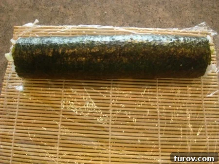 A sheet of nori, filled with rice and other ingredients, being expertly rolled up on a bamboo mat to create a firm sushi cylinder.