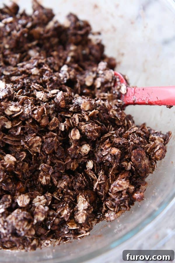 A spatula mixing dark chocolate brownie granola bar ingredients, now coated with syrup, in a large bowl.