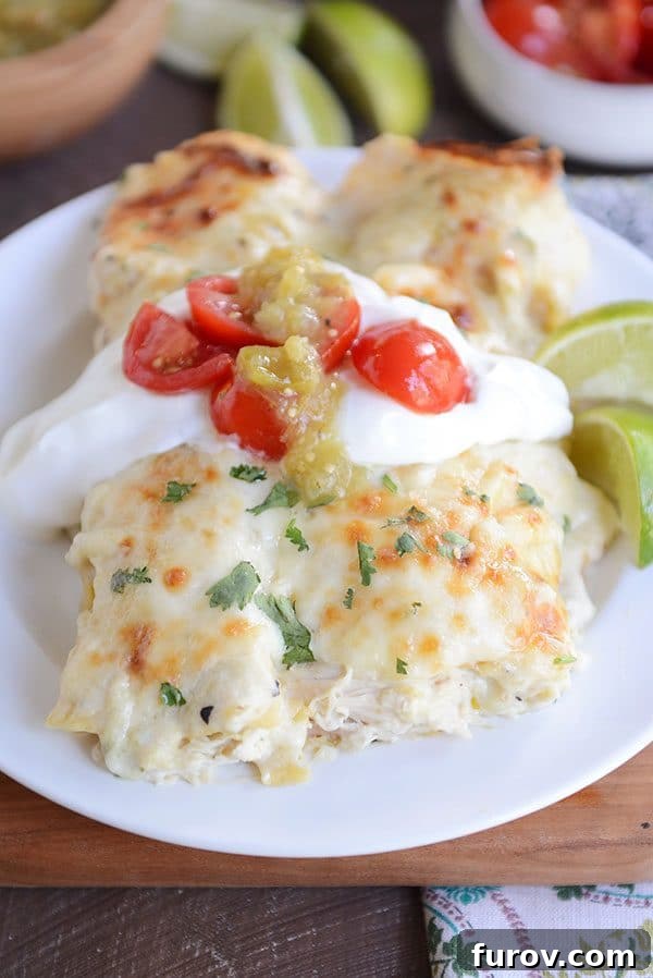 Two baked green chile chicken enchiladas side-by-side on a plate, topped with sour cream and cherry tomatoes and sliced limes on the side.