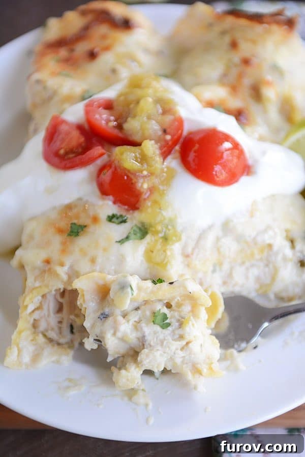 Two baked green chile chicken enchiladas side-by-side on a plate, topped with sour cream and cherry tomatoes and sliced limes on the side.
