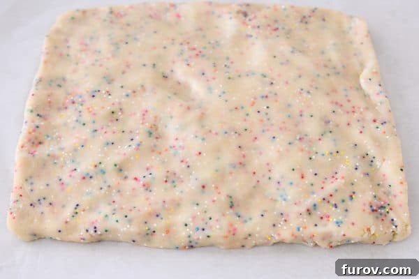Hands gently pressing the funfetti shortbread dough into a smooth rectangle on a baking sheet, preparing it for chilling.
