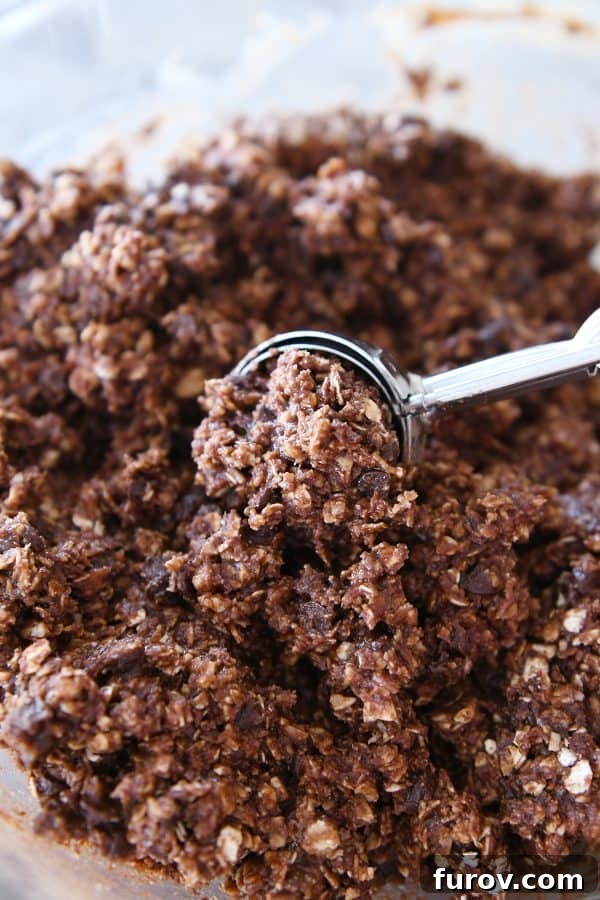 Scooping out uniform portions of chocolate peanut butter granola bite batter onto a parchment-lined baking sheet using a cookie scoop.
