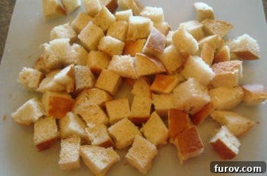 A substantial pile of freshly cut one-inch bread cubes resting on a pristine white cutting board, awaiting seasoning and baking.