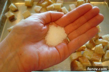 A hand sprinkling coarse salt over a pan of olive oil-coated bread cubes, preparing them for baking into homemade croutons.