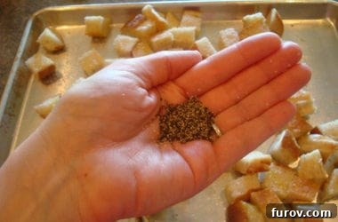 A hand grinding fresh black pepper over a sheet pan filled with seasoned bread cubes, enhancing their flavor before baking.