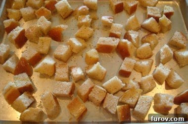 Unbaked, seasoned bread cubes arranged on a metal cookie sheet, perfectly prepared and ready to be placed into the hot oven for toasting.