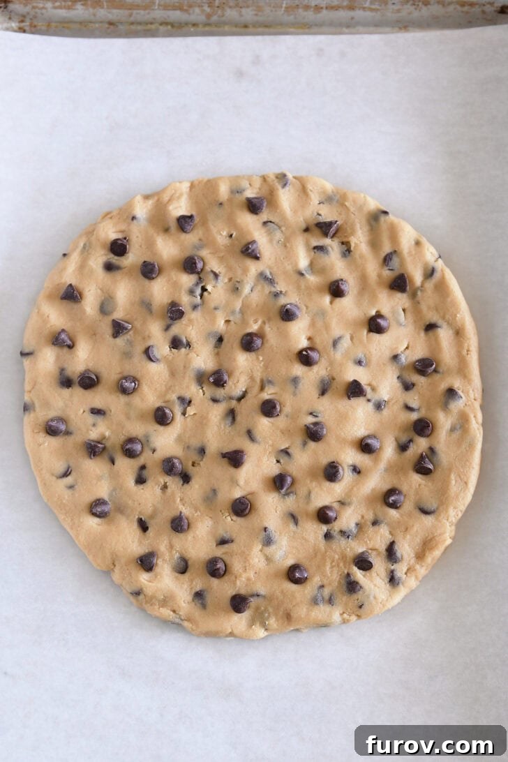 Top-down view of a cookie sheet with parchment paper and a large uncooked chocolate chip cookie dough with extra chocolate chips sprinkled on top in the center.