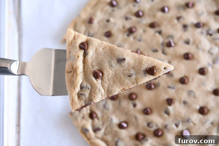 Top down view of a large chocolate chip cookie with a triangle-shaped wedge cut out, being lifted by a metal spatula.