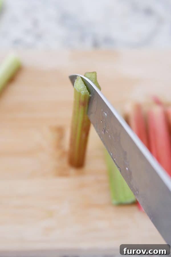 Tangy Rhubarb Custard Delight 8 A hand holds a sharp knife, carefully slicing a rhubarb stalk in half lengthwise on a wooden board, illustrating the preparation technique for thicker rhubarb.
