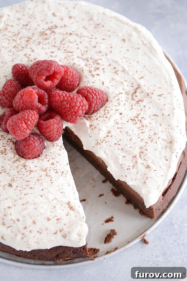 Top view of a decadent chocolate torte covered in whipped cream and fresh raspberries, with a slice removed.