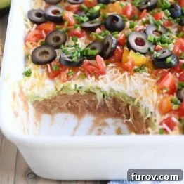 Scoop taken out of 7-layer dip in white casserole dish, showcasing the layers.