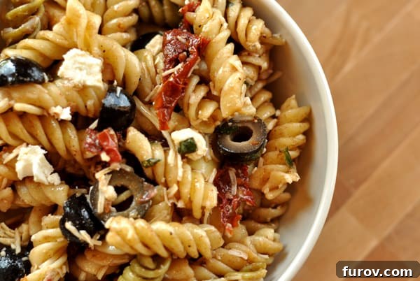 Close-up of rotini pasta with sliced black olives and sun-dried tomatoes in a white bowl, capturing the vibrant ingredients before mixing.