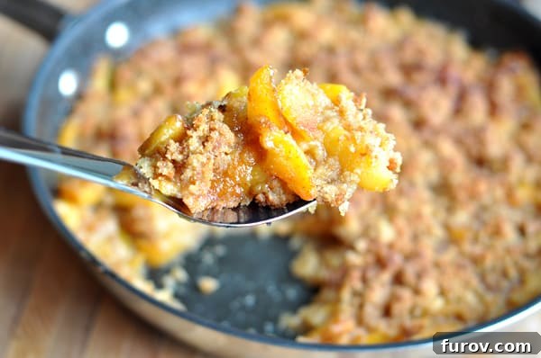 A dark cast-iron skillet filled with bubbling Peach Brown Betty, with a serving spoon scooping out a portion, showing the juicy fruit and crunchy topping.