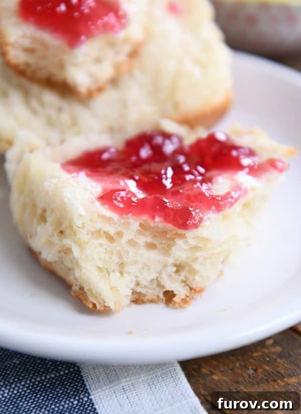 A close-up view of a perfectly fluffy dinner roll, split and adorned with butter and jam, resting on a white plate.