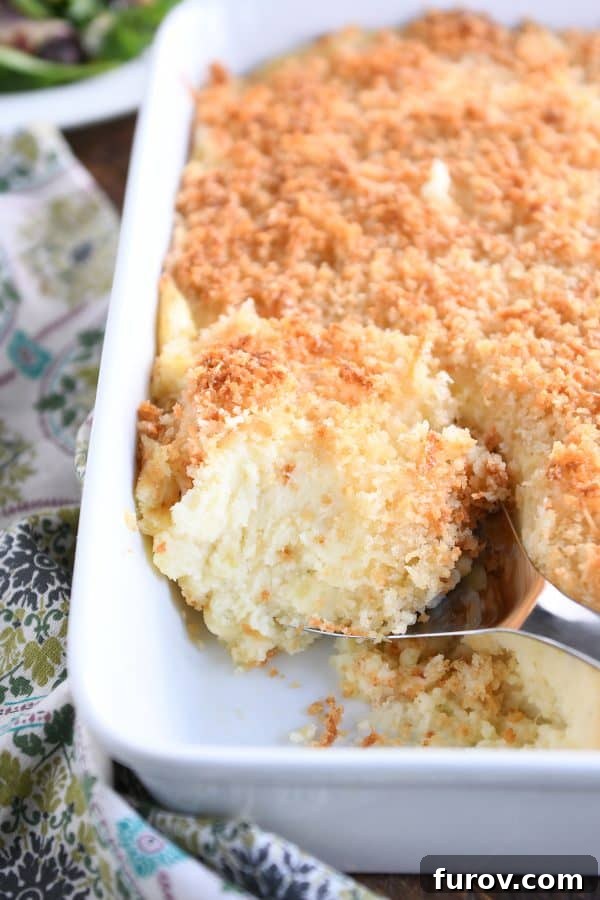 Metal spoon scooping out spoonful of baked mashed potatoes with parmesan topping in white dish.