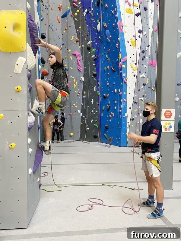 Two teenage boys demonstrate their agility and determination while rock climbing indoors, navigating a tall wall with ropes and safety gear.
