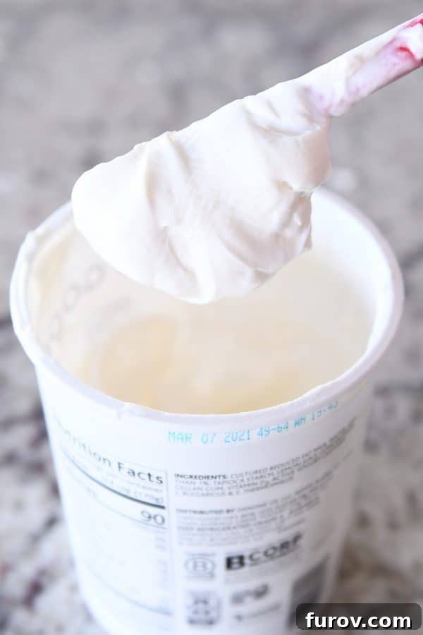 Spatula covered in plain yogurt hovering over a tub of yogurt, showing the thick, scoopable consistency.