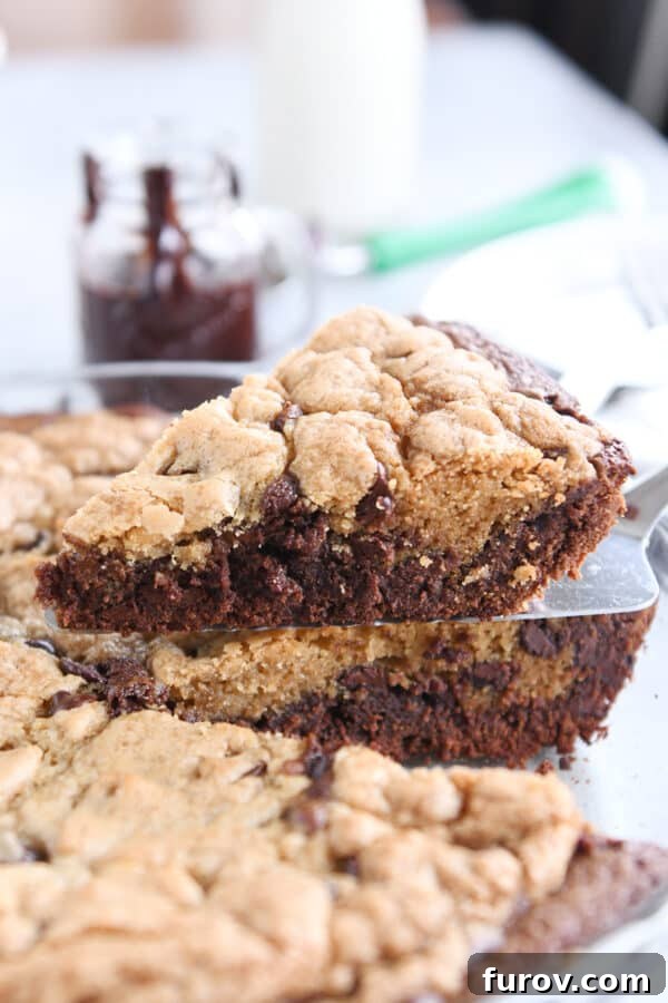 A pie server gracefully lifts a perfectly triangular piece of brookie pie from the glass pie dish, revealing its distinct brownie and cookie layers.