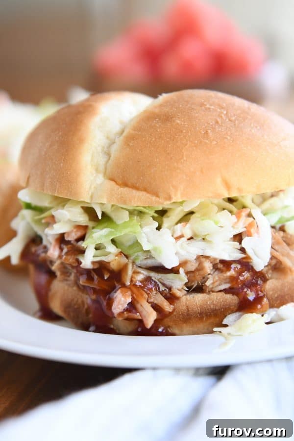 A delectable shredded BBQ pork sandwich, topped with creamy coleslaw, presented on a pristine white plate.