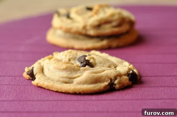 A chocolate chip cookie on a purple table, with two more cookies in the background.