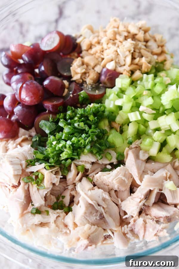 Close-up of shredded chicken, vibrant green onions, crisp celery, juicy grapes, and crunchy nuts in a clear glass bowl, showcasing fresh ingredients.