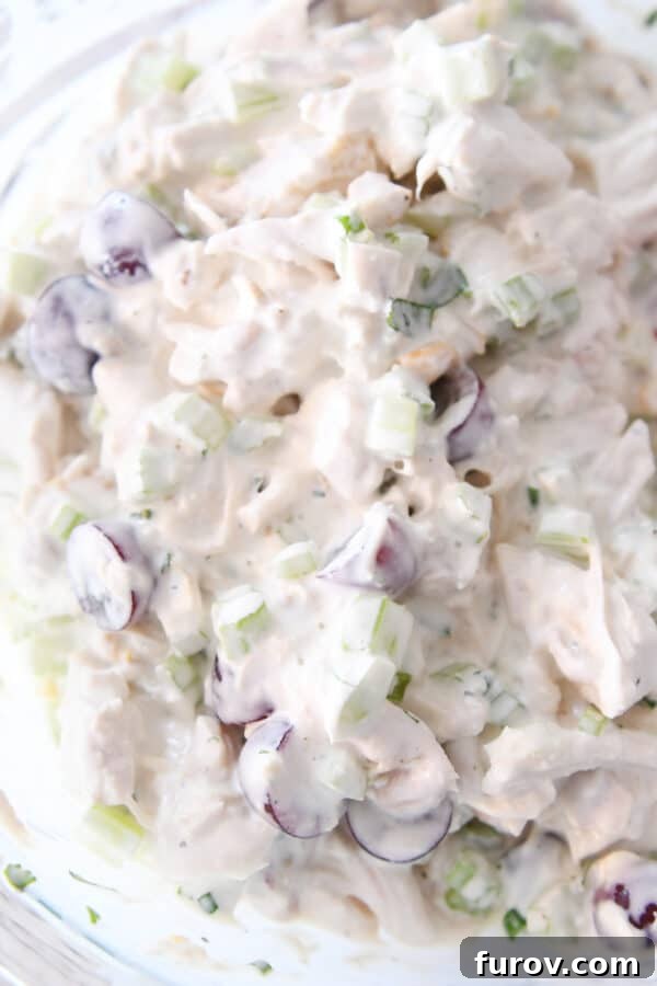 Chicken salad ingredients being mixed with a rich, creamy dressing in a clear glass bowl, highlighting the texture and consistency.