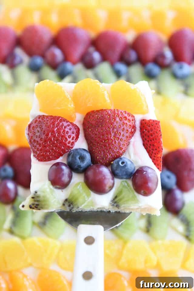 Square of fruit pizza with mandarin oranges, strawberries, blueberries, red grapes and kiwi above sheet pan fruit pizza.