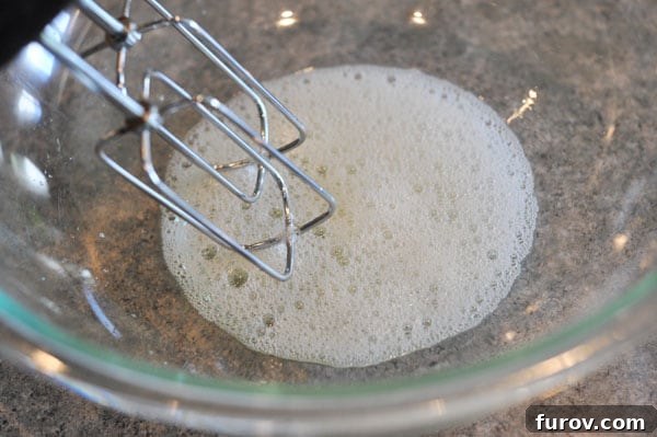 A glass bowl containing egg whites that are becoming frothy as they are whipped by electric beaters.