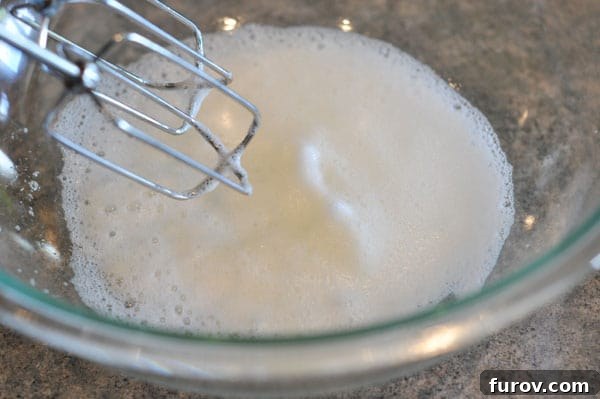 Egg whites being beaten in a glass bowl, now much more foamy and increased in volume.