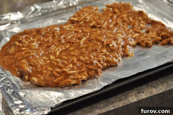Hot caramel and toasted almonds are being spread evenly onto a foil-lined cookie sheet using an offset spatula.