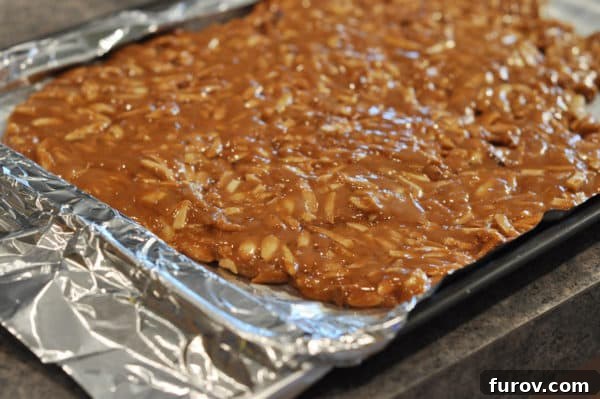 A thin, even layer of caramel-coated almonds is spread across a foil-lined sheet pan, hardening quickly.