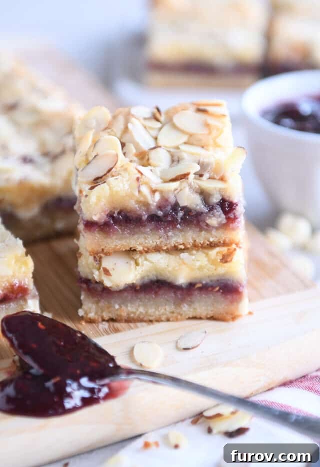 Luscious Raspberry White Chocolate Blondies 6 Two raspberry white chocolate blondie bars stacked on each other on wood cutting board with spoon of raspberry jam.