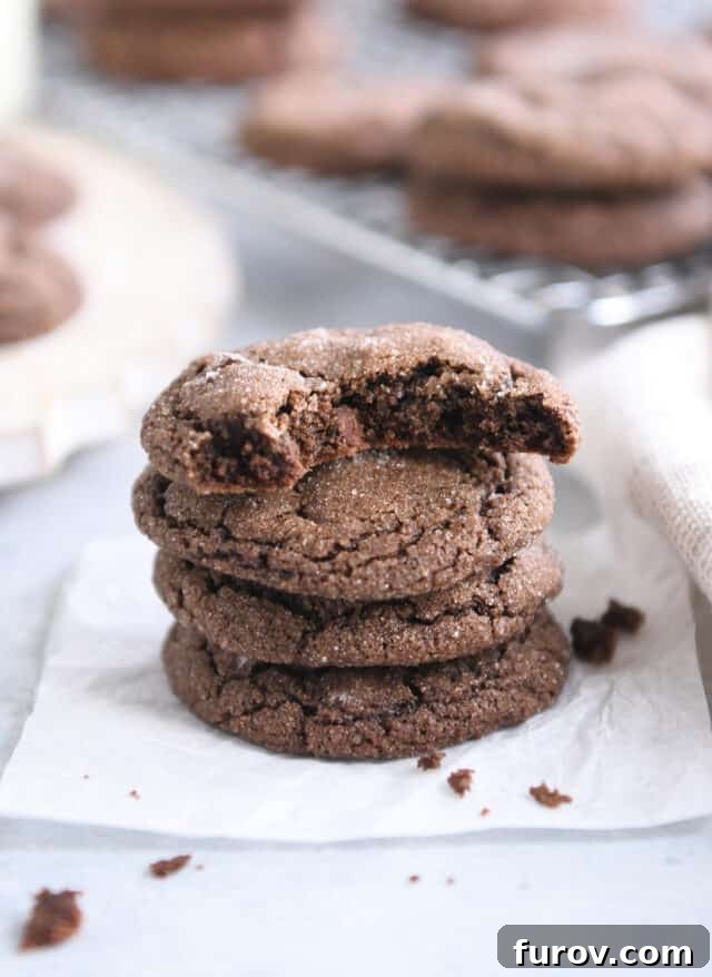 A bite taken out of a chocolate ginger molasses cookie, revealing its soft interior, placed on a stack of three whole cookies.