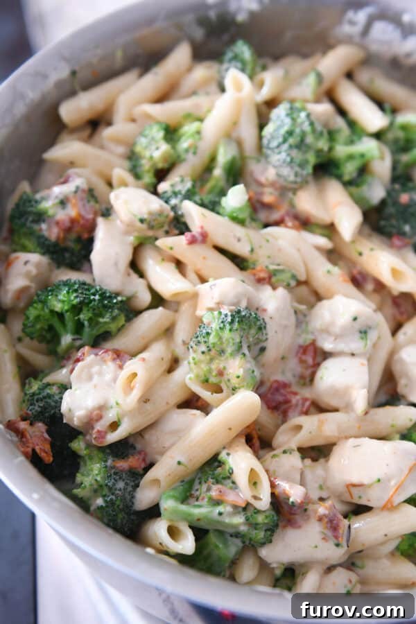 Hearty Chicken and Broccoli Penne Bake with Smoked Mozzarella 5 Close-up of creamy pasta, vibrant broccoli, and sun-dried tomatoes thoroughly mixed in a large stainless steel pot, ready for baking.