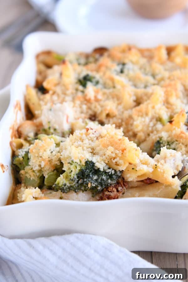 Hearty Chicken and Broccoli Penne Bake with Smoked Mozzarella 7 A serving spoon lifting a generous portion of baked penne, showcasing the chicken, broccoli, melted cheese, and golden-brown breadcrumb topping, from a white casserole dish.