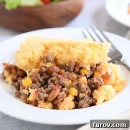 Portion of cornbread topped beef and bean casserole on white plate with fork.