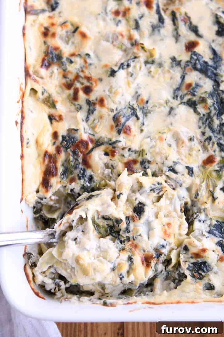 Ultimate Creamy Spinach Artichoke Dip 2 Top down view of a spoon taking a large scoop of spinach artichoke dip out of a white casserole dish full of dip, showcasing its golden, bubbly crust.