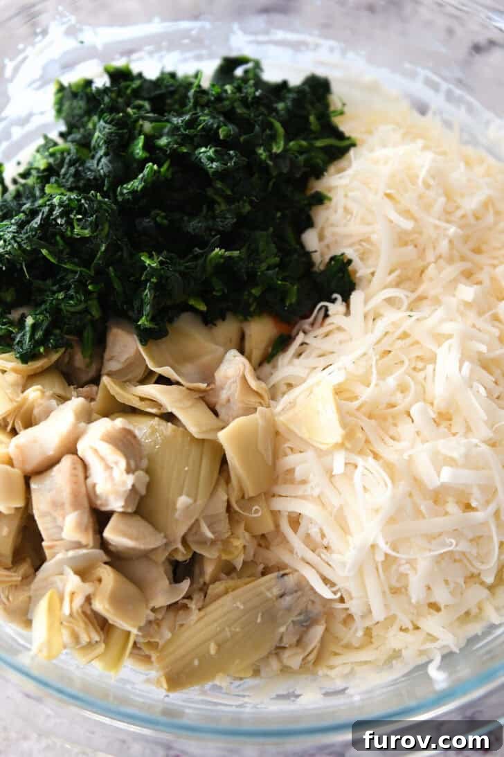 Ultimate Creamy Spinach Artichoke Dip 4 Top down view of a glass bowl with chopped artichokes, shredded cheese, and chopped spinach, combined with the creamy base.